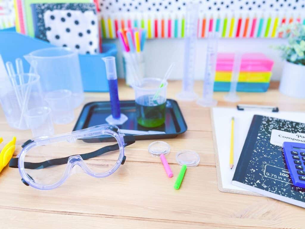 Science resources for the classroom shown on a desk. Magnifying glasses, beakers, a calculator, and safety goggles.